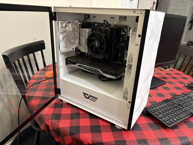 A white computer case with an open glass side panel sits on a table covered in a red and black checkered tablecloth. Inside the case is a motherboard with an "AMD" cooling fan, a "GEFORCE GTX" graphics card, and a white rear exhaust fan. The brand logo "da" is visible on the bottom front panel of the case, and a computer keyboard and monitor are placed on the table nearby.