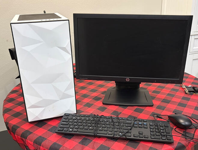 A desktop computer setup on a red and black buffalo plaid tablecloth, featuring a white computer tower with a geometric front panel, a black HP monitor, a wired keyboard, and a wired mouse. Text on the monitor reads, "HP Compaq LA2306x" and "hp".