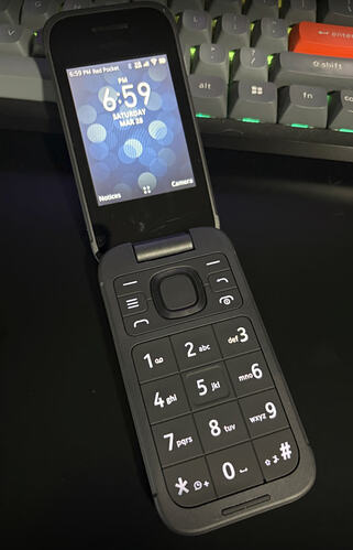 A grey Nokia flip phone is shown in an open position resting on a desk next to a mechanical keyboard. The phone's screen displays the text, "6:59 PM Red Pocket," "6:59," "PM," "SATURDAY MAR 28," "Notices," and "Camera." The device has a standard black alphanumeric keypad.