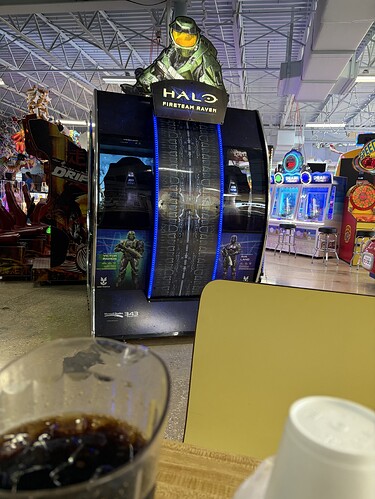 An arcade game featuring "Halo: Fireteam Raven" prominently on a curved black display with blue neon lighting, alongside other arcade games and the roof structure of an arcade; a glass of soda and a white paper cup are present in the foreground. "Halo: Fireteam Raven" is displayed with a large Master Chief helmet on the left, and text is present throughout: "Halo Fireteam Raven,"  "Join Now!". Additional games, including a "Pearl Fishery" game, "Drift" game, and a rotating prize wheel game, are also visible. A yellow table edge is visible in the frame as well.